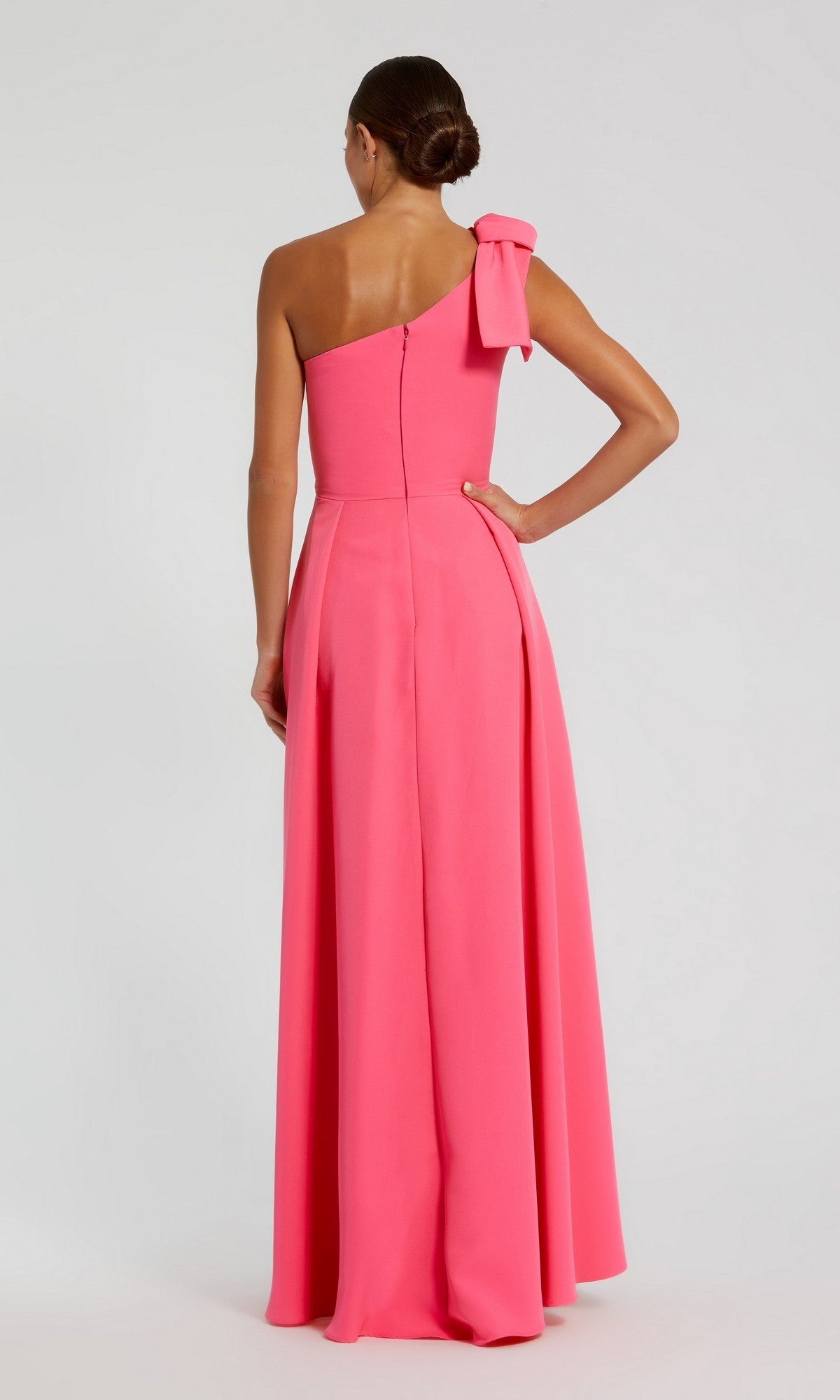 Wearing the Long Formal Dress: Mac Duggal 26989 in bright pink, a woman stands with her back to the camera, showcasing its elegant floor-length silhouette and one-shoulder design accented by a stylish bow detail.